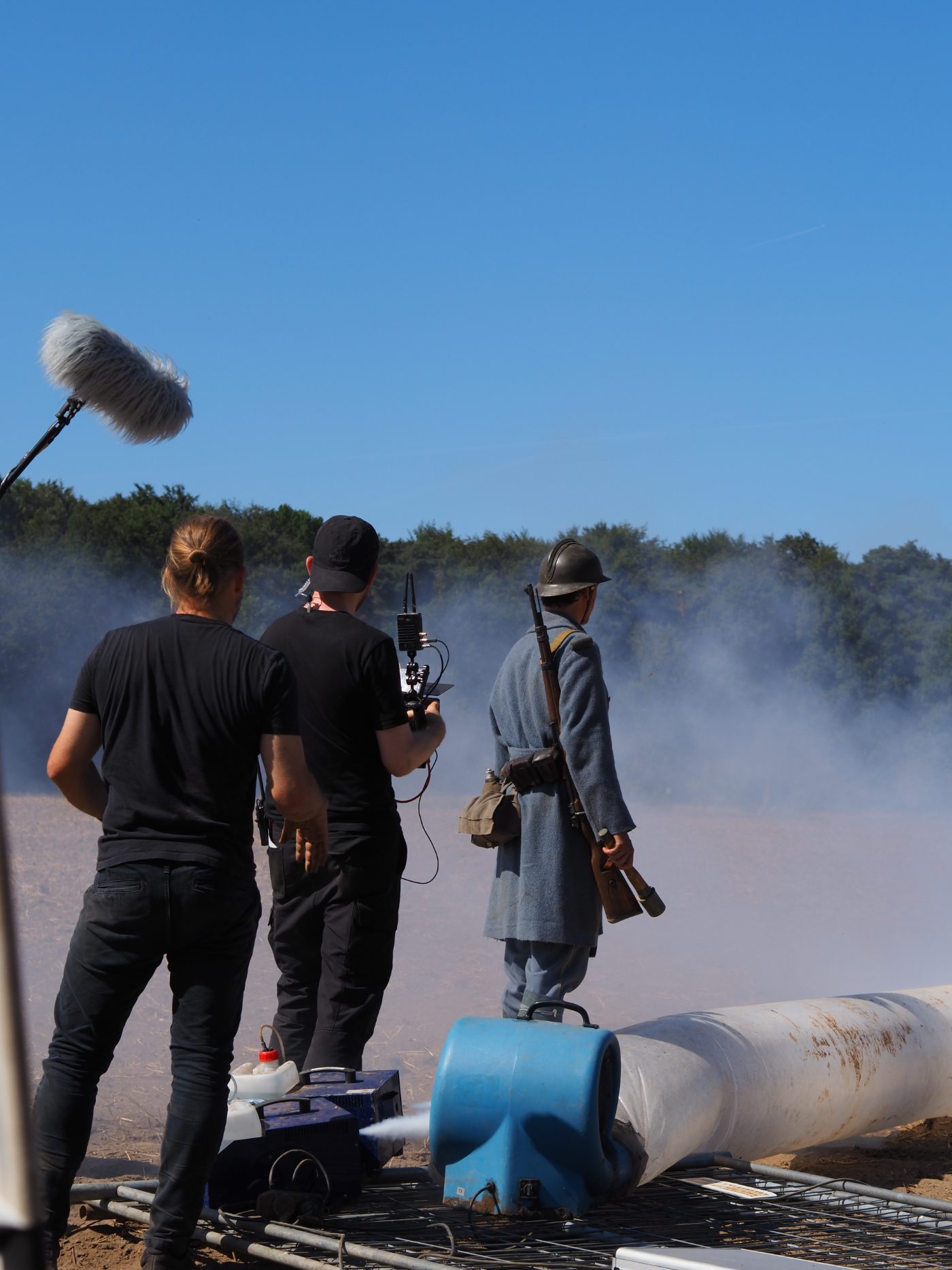 Am Set des Kurzfilms „Graben“, Sommer 2026, fotografiert von Simon Schaugg: zwei Crewmitglieder mit Tonangel und Kamera, daneben ein Darsteller im französischen Militärmantel mit Adrian-Helm und Gewehr, im Rauch über einem Feld.