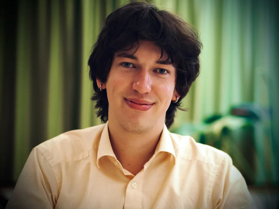 Porträt von Simon Elias Schaugg: lächelnd, mit dunklen halblangen Haaren und hellem Hemd vor einem grünen Vorhang.